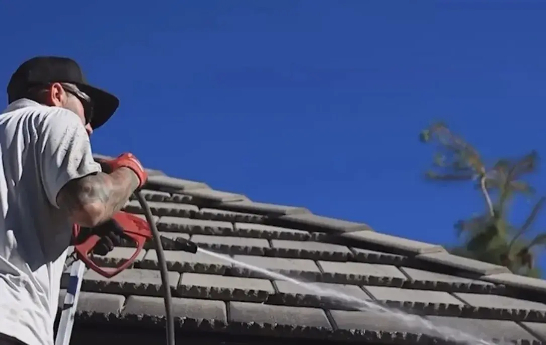 Worker in a black cap and white shirt uses a red pressure washer wand to spray water across shingle roof against blue sky