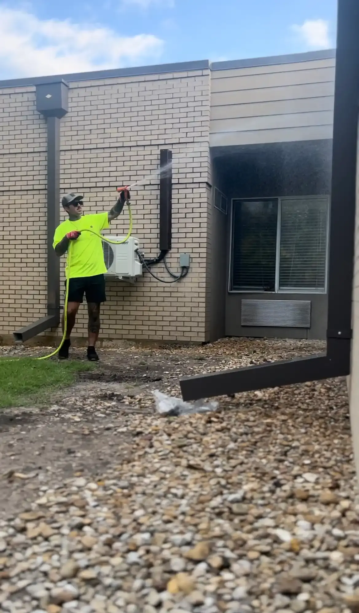 Worker in neon yellow shirt pressure washing a beige brick building wall near an air conditioning unit