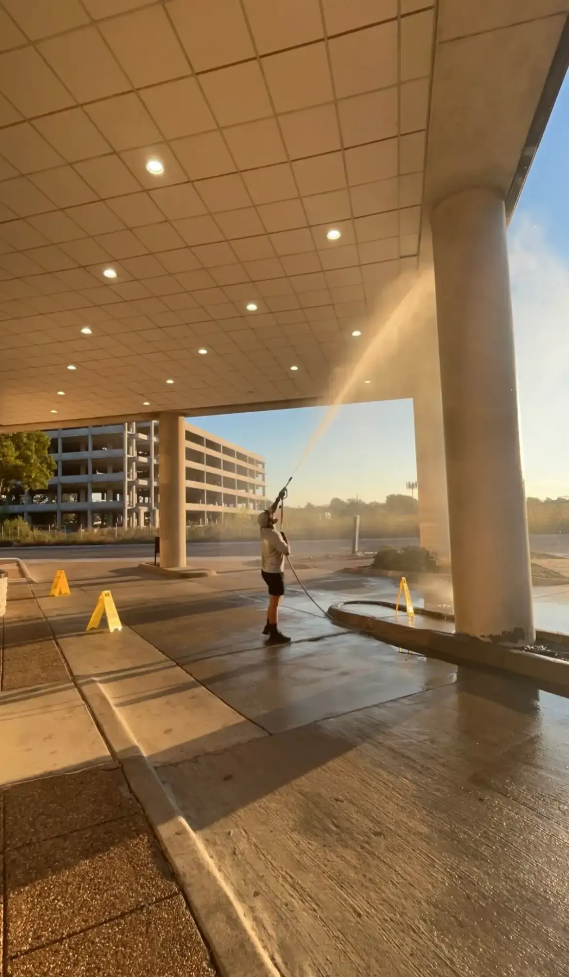Worker pressure-washing a tiled building overhang and columns at a medical center entrance, with yellow caution signs on the