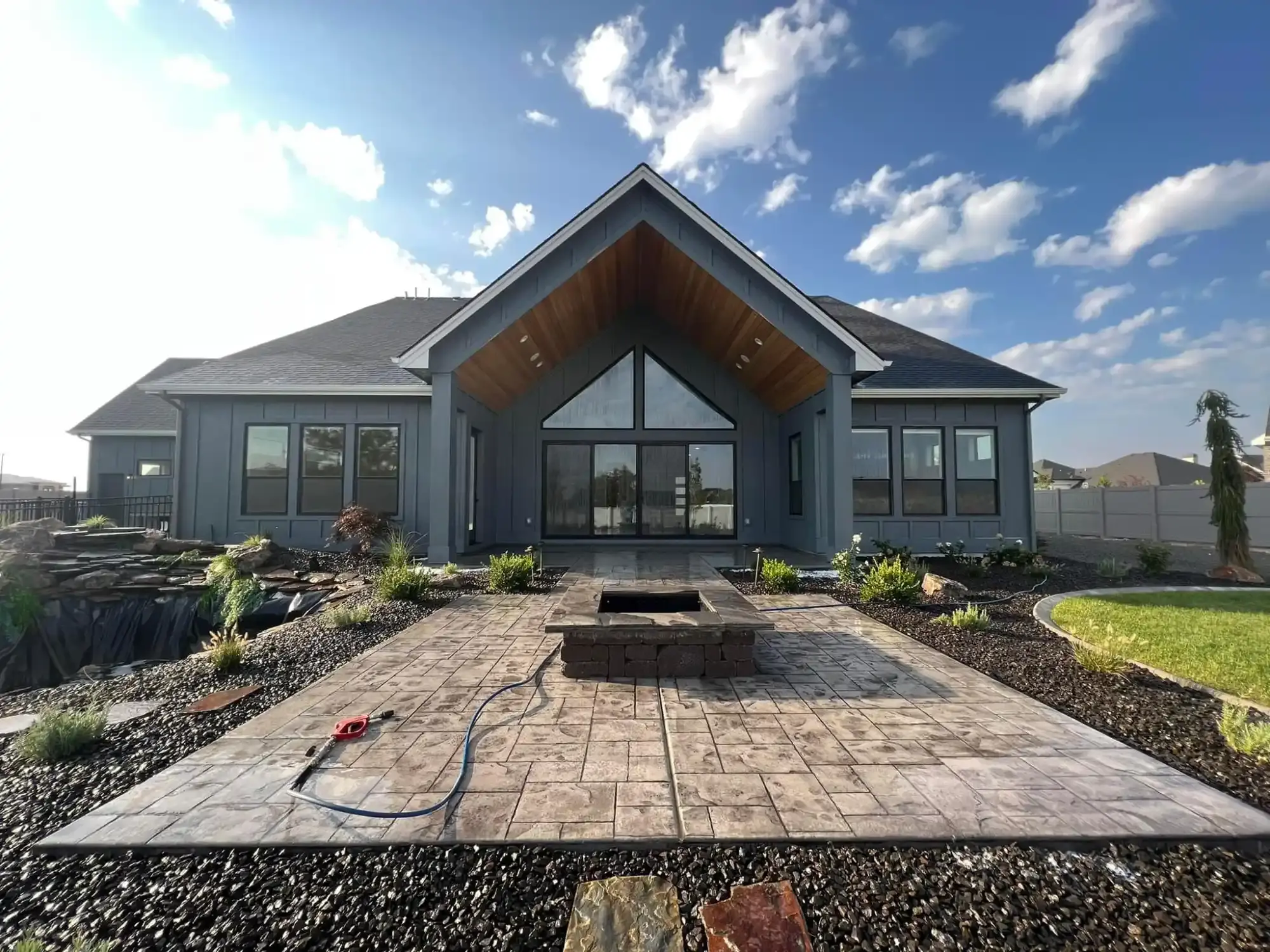Clean stamped concrete patio with a stone fire pit and pressure washer wand in front of a modern gray house