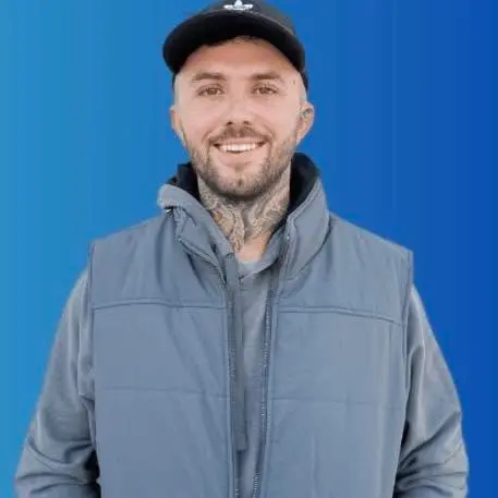 Smiling man with neck tattoos wearing a black cap and gray vest against a blue background