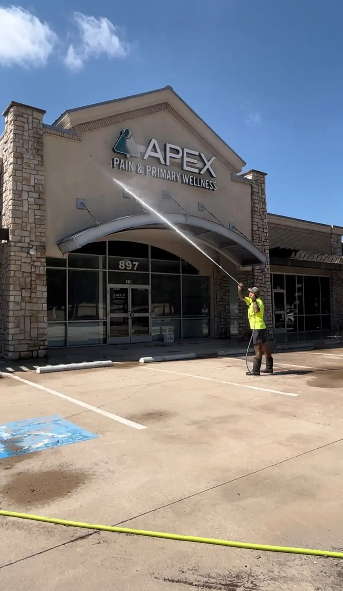 Worker in a high-visibility shirt pressure-washing the facade of Apex Pain & Primary Wellness building